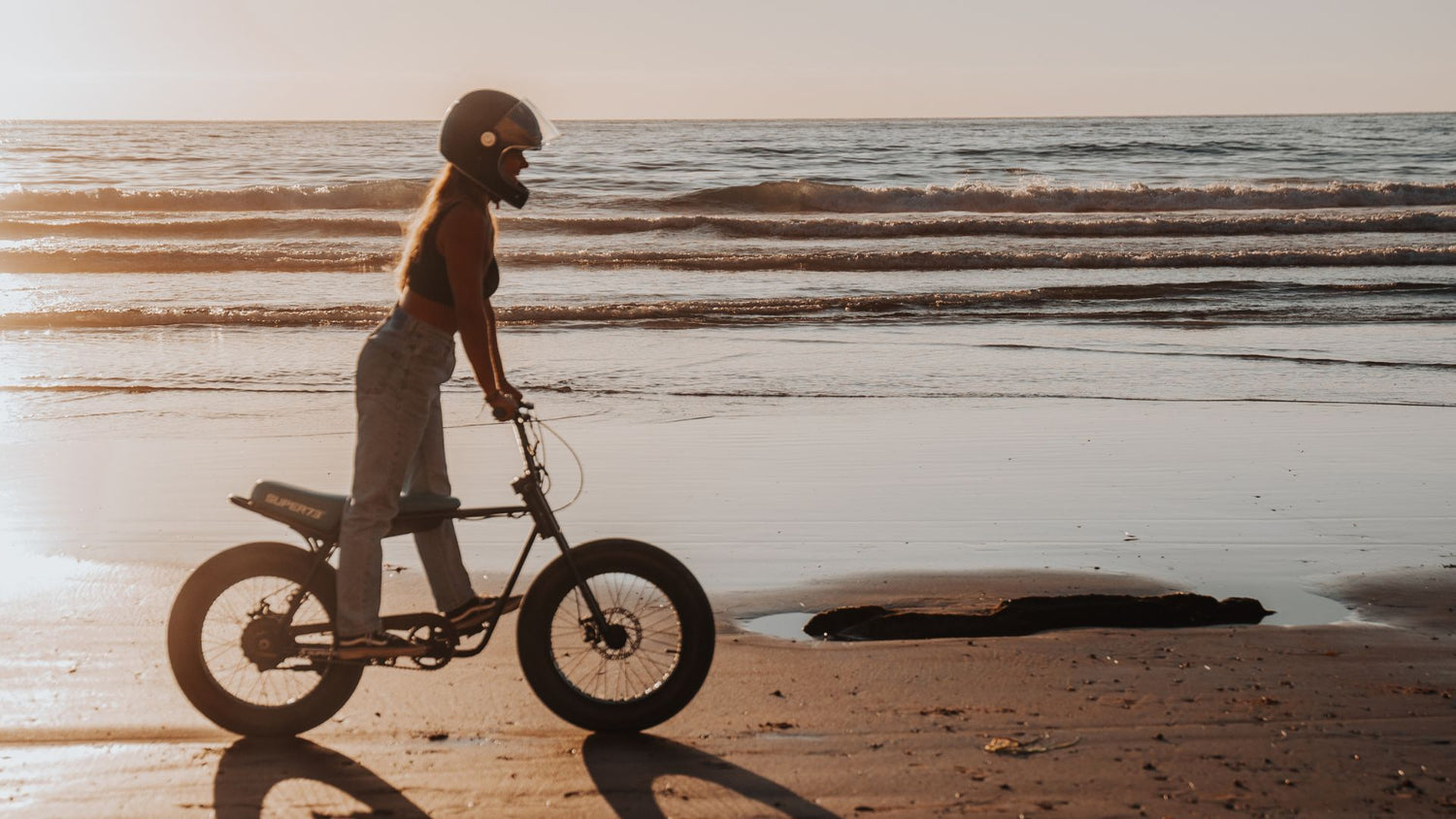 Young woman riding a Super73 ebike with a helmet on across the beach