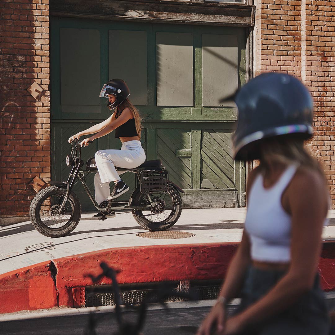 Rider watching another rider riding Super73 S2 ebikes in front of brick building 